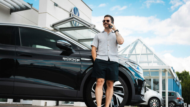 Ein Mann telefoniert entspannt mit einem Smartphone, während er an einen schwarzen VW von Autohaus Socke gelehnt ist.