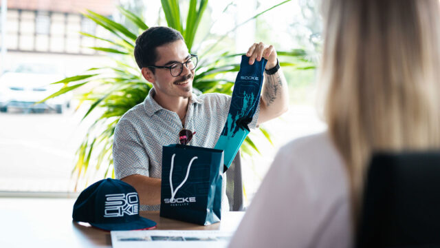 Ein gut gelaunter Mann präsentiert eine blaue "Autohaus Socke"-Socke aus einer passenden Geschenktüte, daneben liegt eine Cap.