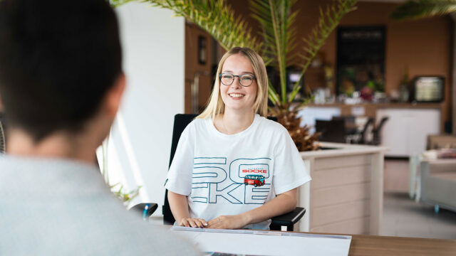 Eine freundliche Mitarbeiterin mit Brille und einem "Autohaus Socke"-T-Shirt lächelt während eines Beratungsgesprächs am Schreibtisch.