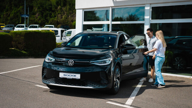 Eine junge Frau öffnet die Fahrertür eines schwarzen VW von Autohaus Socke Flexirent, während ein Mann daneben steht.