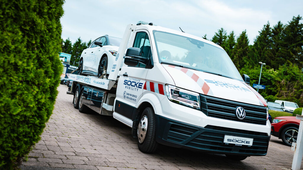 Ein weißer Abschleppwagen von „AUTOHAUS SOCKE MOBILITY“ steht auf einem gepflasterten Hof. Auf seiner Ladefläche transportiert er ein weißes Auto. Links im Bild befindet sich eine grüne Hecke.