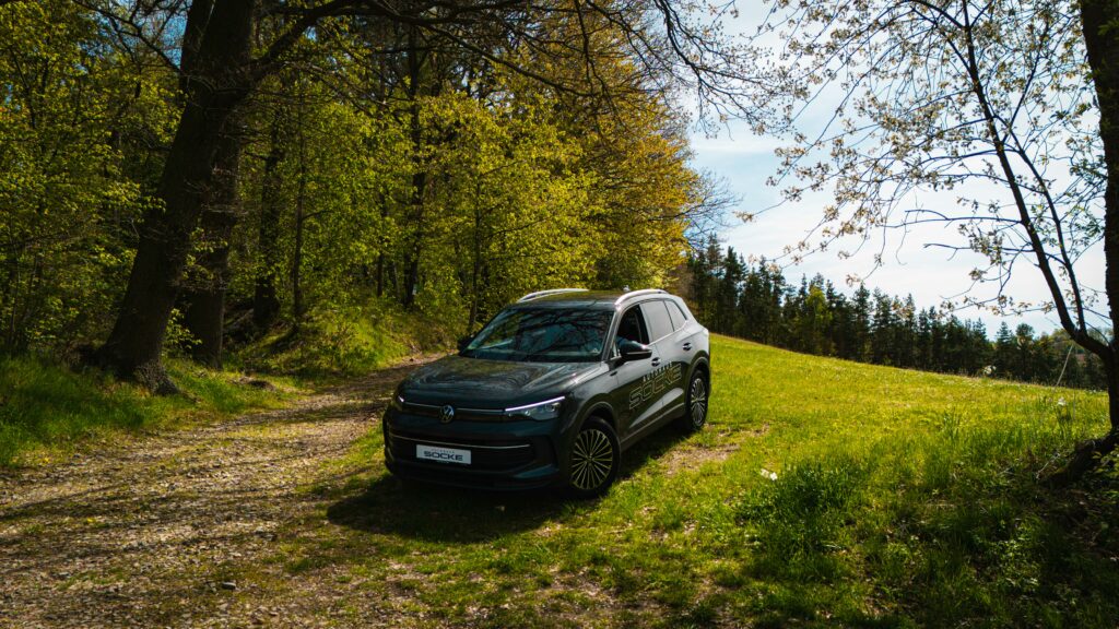 Ein dunkelgrauer VW Tiguan mit der seitlichen Aufschrift „Autohaus Socke“ steht auf einer leicht ansteigenden Wiese am Rande eines Frühlingswaldes unter hohen Bäumen.