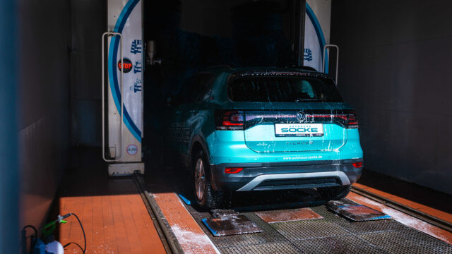 Ein türkisfarbener Volkswagen T-Cross fährt in einer automatischen Waschanlage durch die rotierenden blauen Bürsten. Das Heck des Autos wird mit Wasser abgespritzt.
