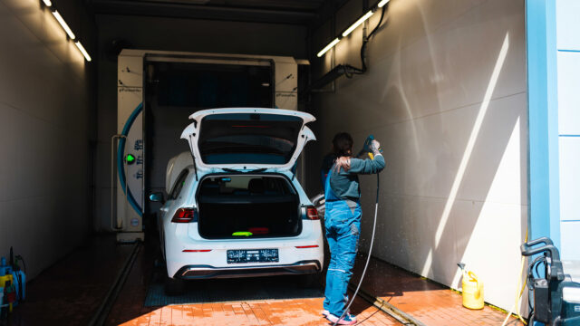 Eine Person in blauer Arbeitskleidung steht in einer Waschhalle und reinigt ein weißes Auto mit einem Hochdruckreiniger. Die Heckklappe des Wagens ist weit geöffnet, im Hintergrund ist die Einfahrt zu einer automatischen Waschanlage zu sehen.