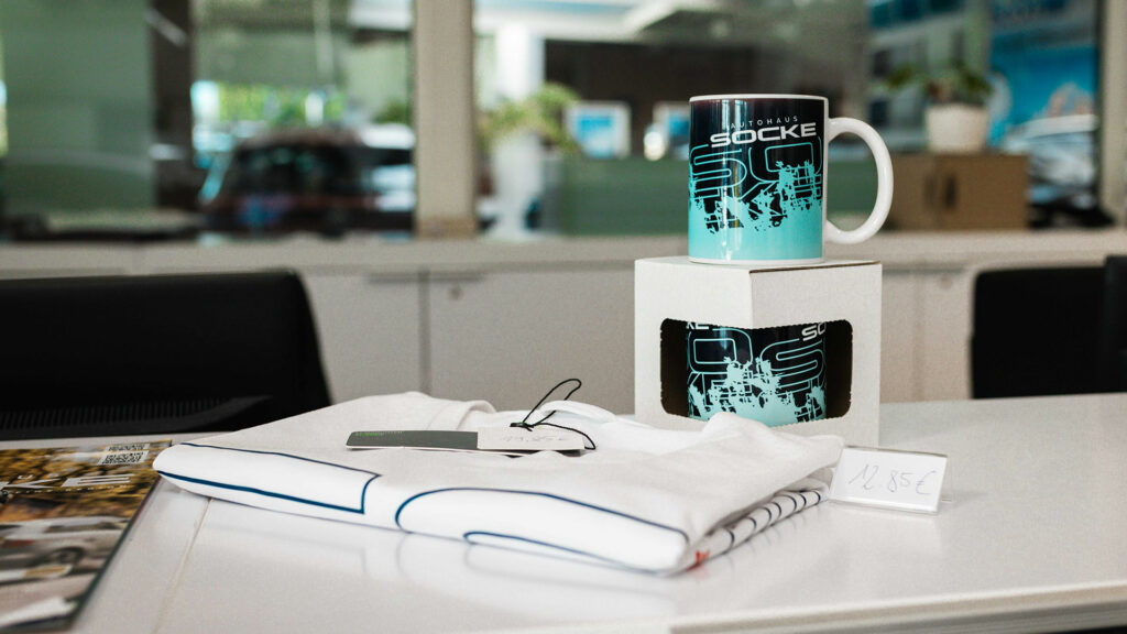 Merchandiseartikel von Autohaus Socke liegen auf der weißen Rezeption. Ein weißes TShirt mit dem Logo von Autohaus Socke und ene Tasse mit Hellblauem Logo im coolen Grafittistil