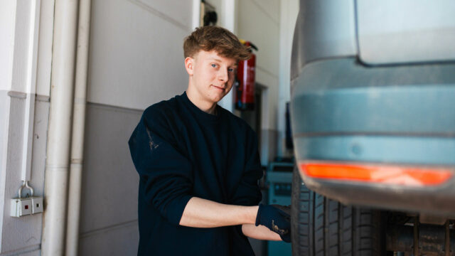 Ein junger Mechaniker im dunklen Pullover schaut lächelnd in die Kamera, während er an dem Reifen eines Autos arbeitet, das sich auf einer Hebebühne befindet.