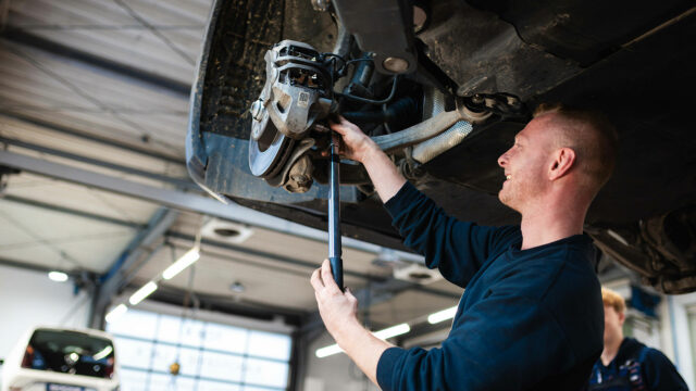 Ein lächelnder Automechaniker arbeitet in einer hellen Werkstatt unter einem angehobenen Fahrzeug und zieht mit einem langen Drehmomentschlüssel Schrauben an der Radaufhängung und Bremsscheibe fest.