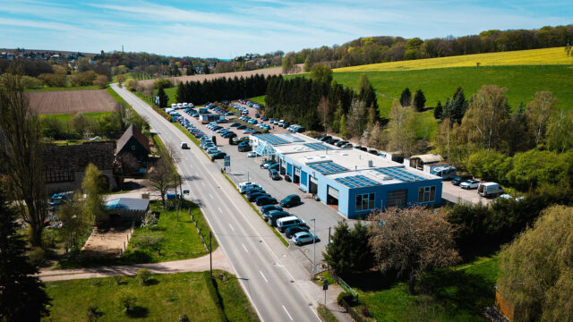 Luftaufnahme des langgestreckten, hellblauen Autohaus-Socke-Gebäudes mit Solarmodulen auf dem Dach. Neben einer vorbeiführenden Landstraße sind zahlreiche geparkte Fahrzeuge und die umliegende Hügellandschaft sichtbar.