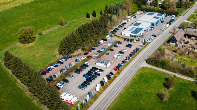 Drohnen-Vogelperspektive auf das gesamte Gelände vom Autohaus Socke, eingebettet in eine grüne Landschaft mit Rapsfeldern. Zu sehen sind das Hauptgebäude mit Solaranlagen und ein großer, gut gefüllter Kunden- und Gebrauchtwagenparkplatz.
