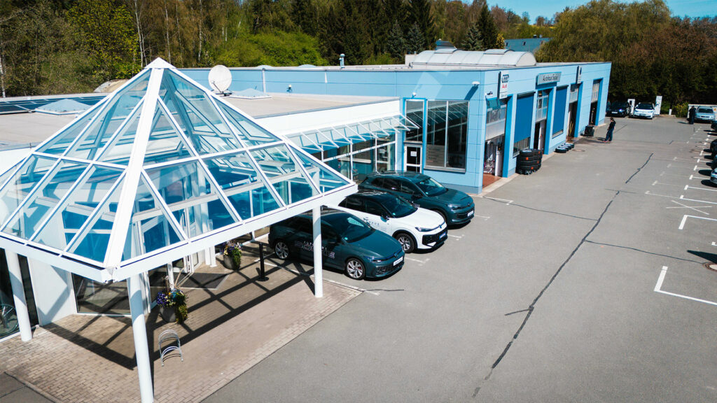 Drei moderne Volkswagen-Modelle (zwei dunkelgraue, ein weißes) stehen ordentlich aufgereiht vor dem hellblauen Werkstatt- und Servicegebäude des Autohauses Socke unter einem Glasvordach.