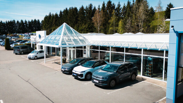 Die gläserne, pyramidenförmige Eingangshalle des Autohauses Socke mit dem markanten VW-Logo im Hintergrund. Davor stehen verschiedene Neuwagen auf dem sauberen Hof.