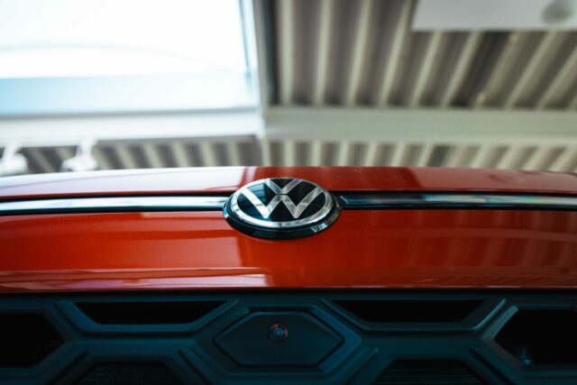 Detailaufnahme des verchromten Volkswagen-Logos auf der roten Motorhaube eines VW T-Roc, direkt über dem schwarzen Kühlergrill.