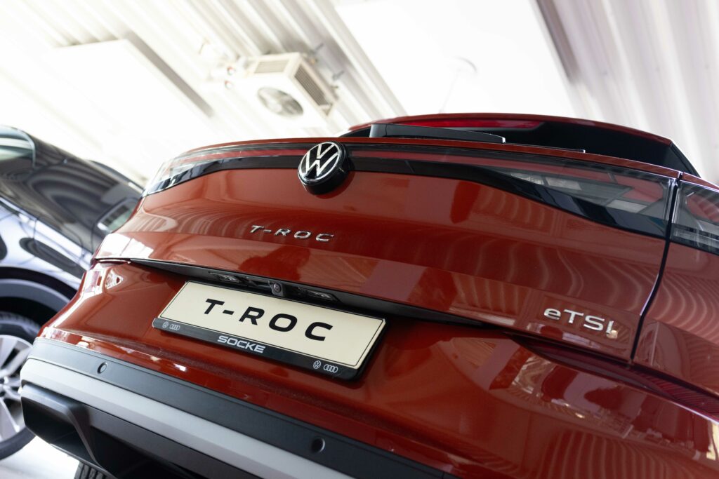 Nahaufnahme des Hecks eines roten VW T-Roc mit dem VW-Logo, dem verchromten Modellschriftzug und dem „T-ROC“-Kennzeichen.