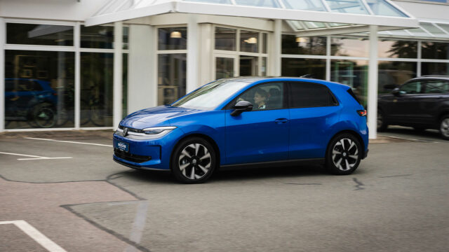 Seitliche Fahrtaufnahme eines blauen Volkswagen ID. POLO mit Fahrerin auf einem Firmengelände vor einer Glasfront.
