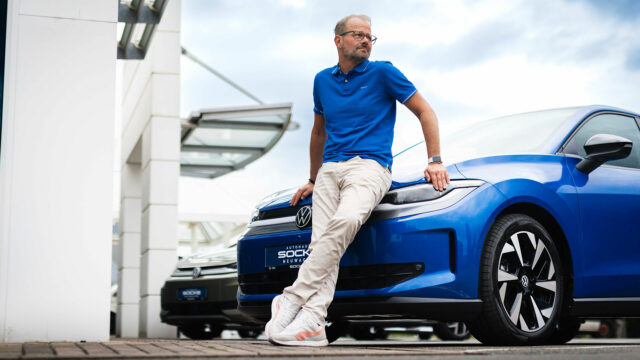 Mann in blauem Poloshirt und heller Hose lehnt entspannt an der Front eines blauen Volkswagen ID. POLO.