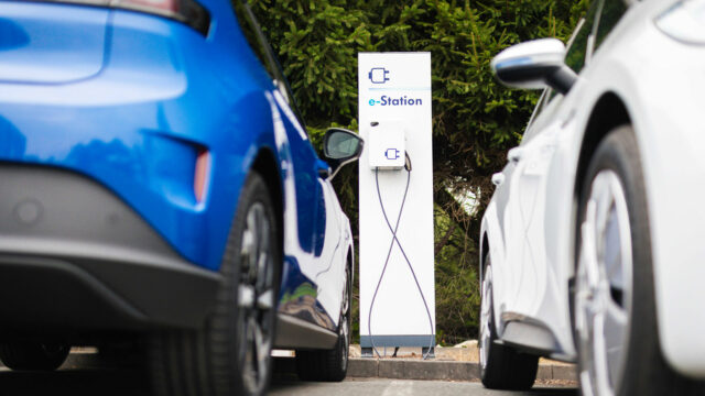 Weiß-blaue Ladestation (e-Station) mit zwei angeschlossenen Ladekabeln, eingerahmt von zwei Elektrofahrzeugen.