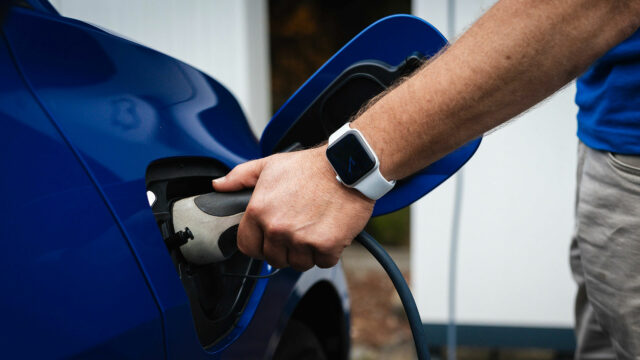 Nahaufnahme einer Hand mit Smartwatch, die einen Ladestecker in den Ladeanschluss eines blauen Elektrofahrzeugs steckt.