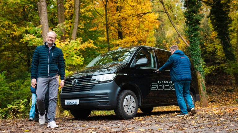 Mitarbeiter von Autohaus Socke steht neben einenm schwarzen VW Bulli inmitten eines Herbstwaldes